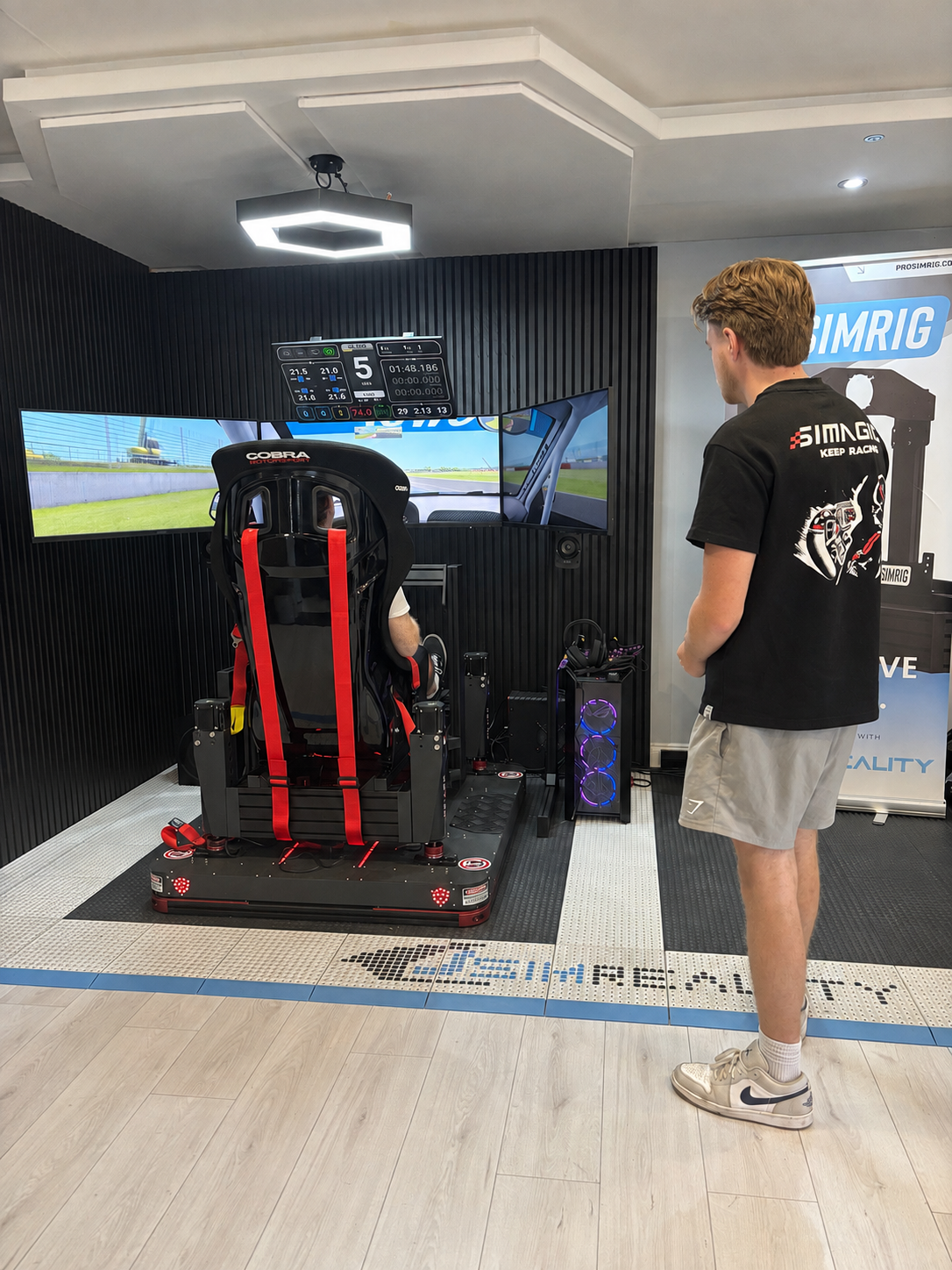 A man wearing a black t-shirt and gray shorts stands next to a racing simulator setup in a home theater or simulator room. The simulator features a black and red racing seat, three screens showing a virtual race, and a computer tower.